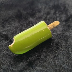 Könnte beinhalten: Ein grüner Eis am Stiel mit einem goldenen Stiel. Das Eis am Stiel liegt auf einer schwarzen Oberfläche.
