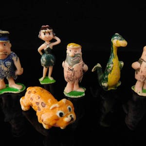 May include: A collection of vintage Flintstones character figurines. The figures include Fred Flintstone, Wilma Flintstone, Barney Rubble, Dino, and others. The figurines are made of plastic and have bright colors. The figures are standing on green bases.