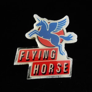 Könnte beinhalten: Vintage Anstecknadel mit einem blauen geflügelten Pferd-Emblem über einem roten Kreis. Die Worte "FLYING HORSE" sind in weißen Buchstaben auf rotem rechteckigem Hintergrund mit silbernem Rand dargestellt. Die Nadel ist vor schwarzem Hintergrund.