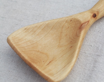 May include: A light-colored wooden kitchen utensil with a wide, triangular-shaped head and a smooth, rounded handle. The wood grain is visible throughout the surface, showing natural patterns and small knots in the material.
