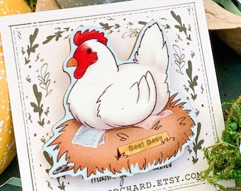 May include: A white chicken with a red comb sits in a nest made of straw. The chicken is holding a sign that says "Best Bess". The image is on a white card with a brown border and green leaves around the edges. The card is sitting on a table with a green moss background.