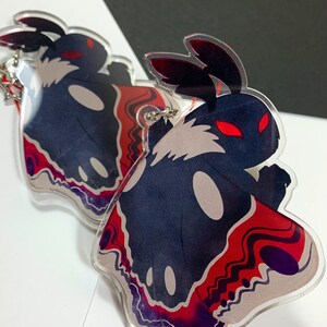 May include: Two black and white moth-shaped acrylic charms with red and purple accents. The charms have red eyes and are attached to silver keychains.