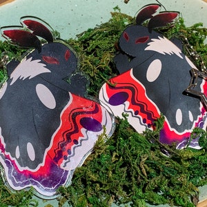 May include: Two black and white moth-shaped acrylic charms with red and purple wings. The charms have a star-shaped metal charm attached to them.