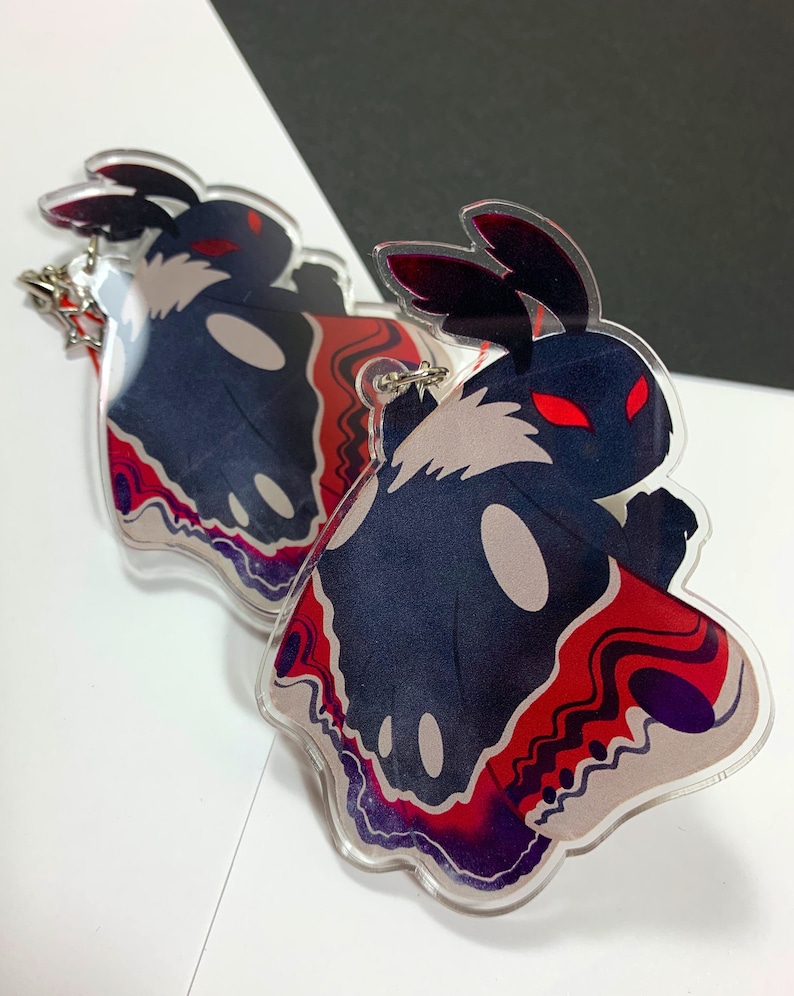 May include: Two black and white moth-shaped acrylic charms with red and purple accents. The charms have red eyes and are attached to silver keychains.