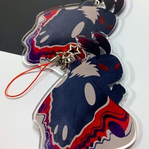 May include: Two acrylic charms featuring a black and white moth with red eyes and a red, purple, and white patterned design. The charms are attached to red string keychains.