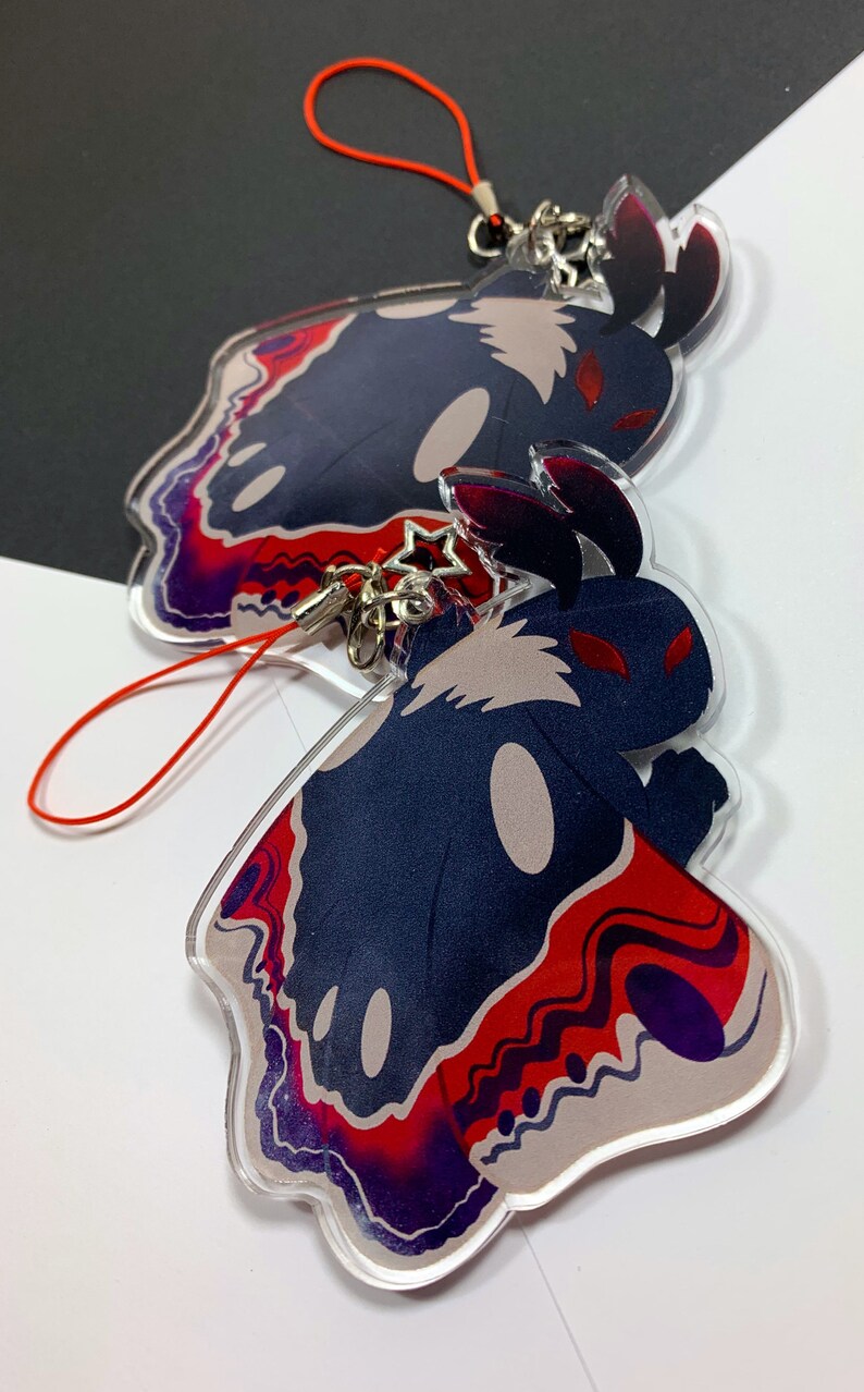 May include: Two acrylic charms featuring a black and white moth with red eyes and a red, purple, and white patterned design. The charms are attached to red string keychains.