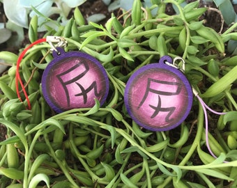 May include: Two round wooden charms with purple and white paint. The charms feature a Japanese kanji character. The charms are attached to red and purple strings.