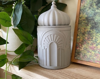 May include: A matte light gray cylindrical container with a removable lid featuring a dome-shaped top. The exterior of the container is decorated with embossed architectural arch motifs that wrap around the entire circumference of the vessel.