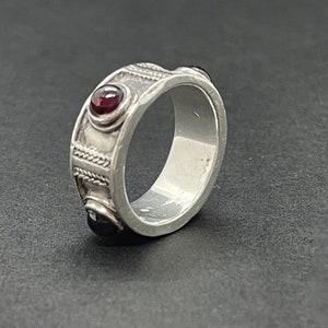 Peut inclure: Une bague en argent avec une bande texturée et trois pierres précieuses rouges. La bague est posée sur un fond noir.
