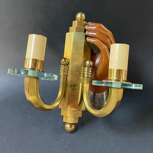 May include: Vintage brass wall sconce with two candle holders. The sconce features a stylized design with a wooden hand and clear glass accents. The sconce has a gold finish and is mounted on a dark background.