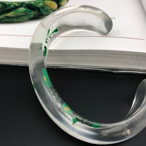 May include: A clear plastic bangle bracelet with a green floral design. The bracelet is thick and has a rounded shape.