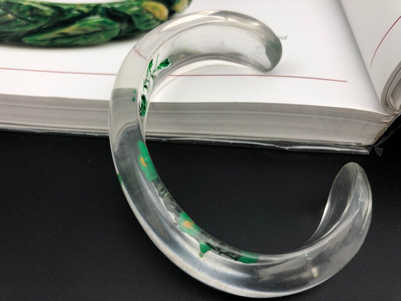 May include: A clear plastic bangle bracelet with a green floral design. The bracelet is thick and has a rounded shape.