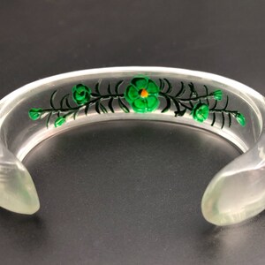 May include: A clear plastic bangle bracelet with a green floral design. The flowers have yellow centers.