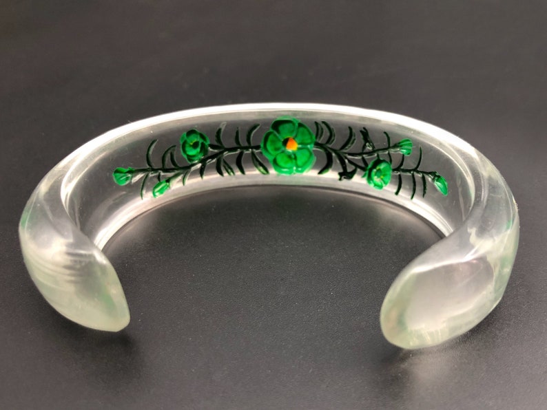 May include: A clear plastic bangle bracelet with a green floral design. The flowers have yellow centers.