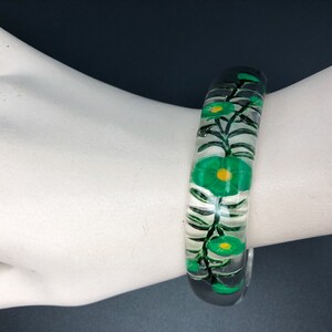 May include: Clear plastic bangle bracelet with a green floral design. The flowers have yellow centers and are painted on the inside of the bangle.