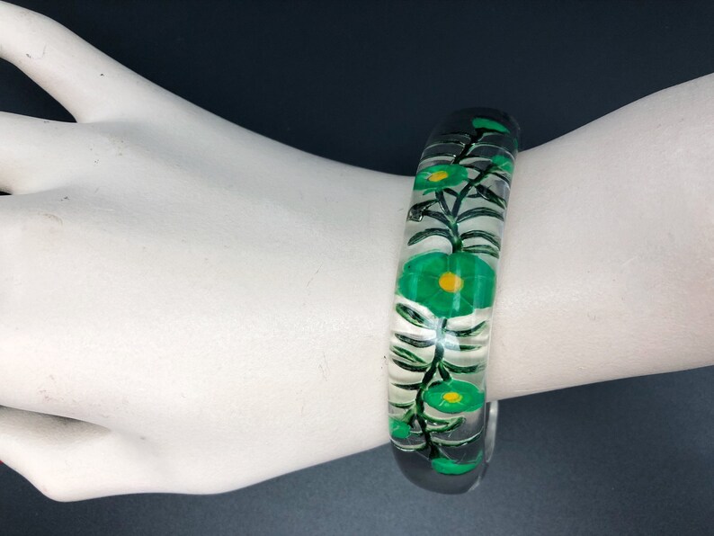 May include: Clear plastic bangle bracelet with a green floral design. The flowers have yellow centers and are painted on the inside of the bangle.