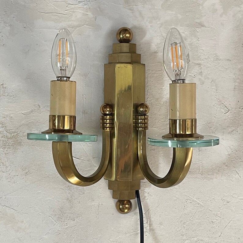 May include: A brass Art Deco wall sconce with two light fittings. Each fitting has a clear glass bulb, a beige candle sleeve, and a round, clear glass plate. The sconce has a geometric design with a central column and curved arms.