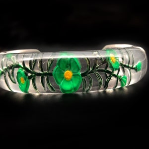 May include: A clear glass bangle bracelet with a green floral design. The flowers have yellow centers and are painted on the inside of the bracelet.