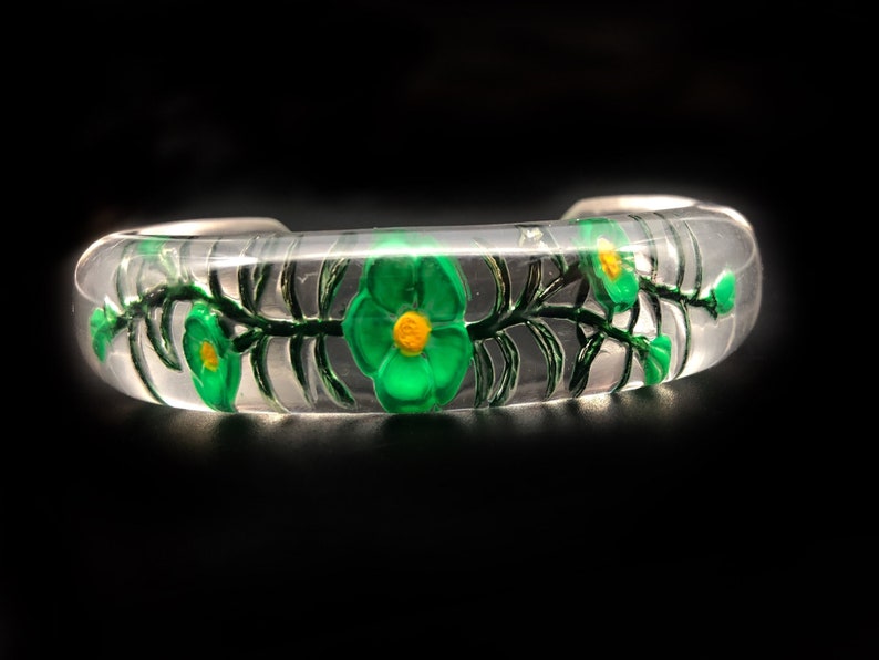 May include: A clear glass bangle bracelet with a green floral design. The flowers have yellow centers and are painted on the inside of the bracelet.