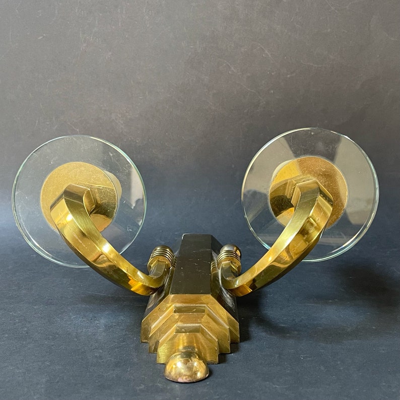 May include: A vintage brass wall sconce with two clear glass disc shades. The sconce has a geometric design with curved arms and a central black accent. The design is Art Deco inspired.