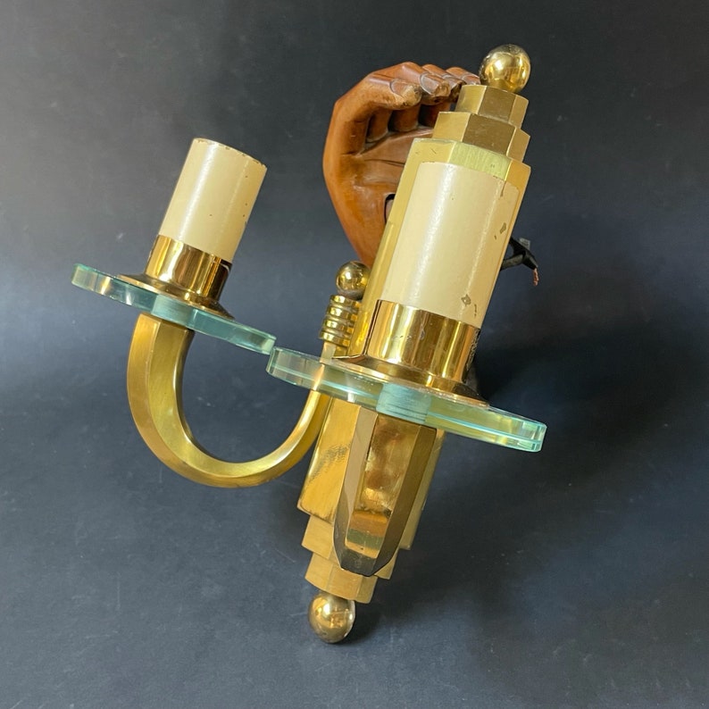 May include: Art Deco wall sconce in brass, with two off-white cylindrical light fittings. The sconce has clear glass details and a curved design. The fixture is mounted on a wooden hand sculpture.