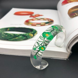 May include: A clear plastic bangle bracelet with a green floral design. The design features green leaves and yellow flowers.