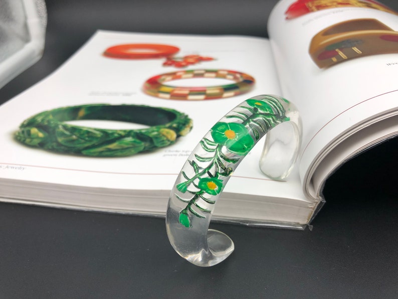 May include: A clear plastic bangle bracelet with a green floral design. The design features green leaves and yellow flowers.