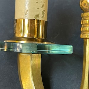 May include: Close-up of a brass candle holder with a clear, circular glass accent. The candle holder has a gold finish and a curved design. A partially burned candle is in the holder.