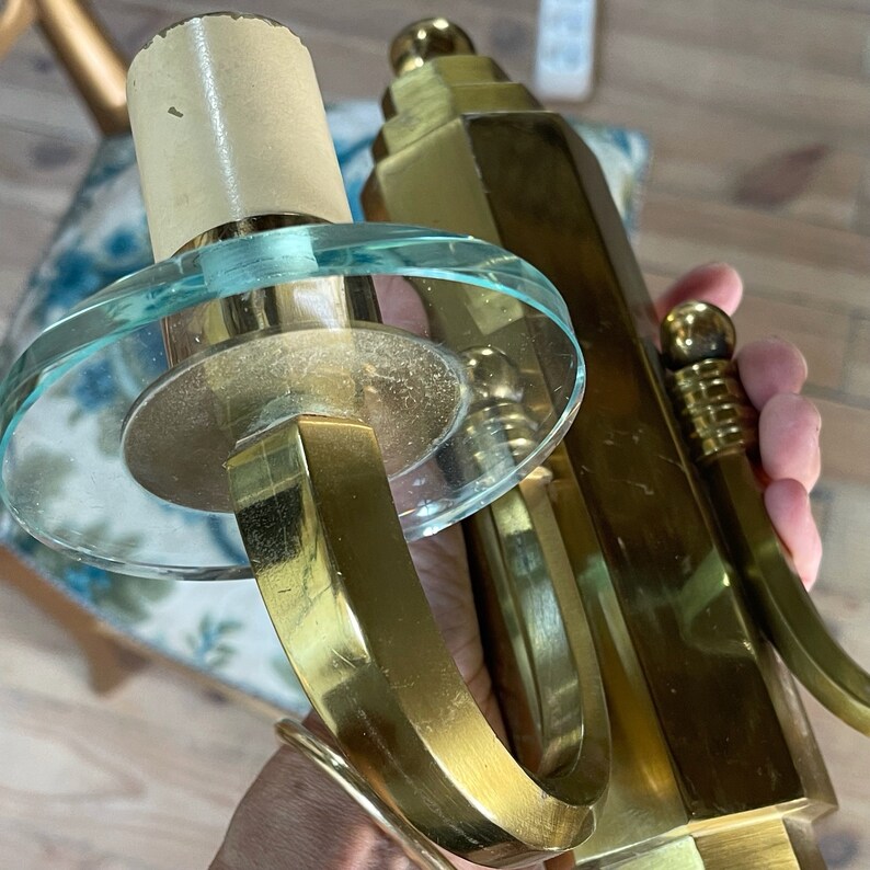 May include: Two vintage brass sconces with clear glass details. One sconce has a circular glass shade and a curved arm. The other has a stepped, geometric design. The sconces are held in a hand.