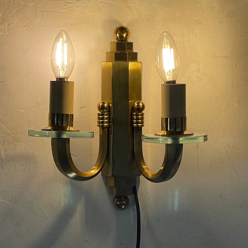 May include: A vintage brass wall sconce with two light bulbs. The sconce has a geometric design with clear glass accents and a black power cord. The bulbs are lit, casting a warm glow.