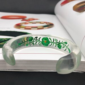 May include: A clear plastic bangle bracelet with a green floral design painted on the inside. The design features a vine with leaves and flowers.