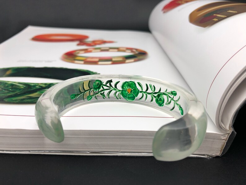 May include: A clear plastic bangle bracelet with a green floral design painted on the inside. The design features a vine with leaves and flowers.