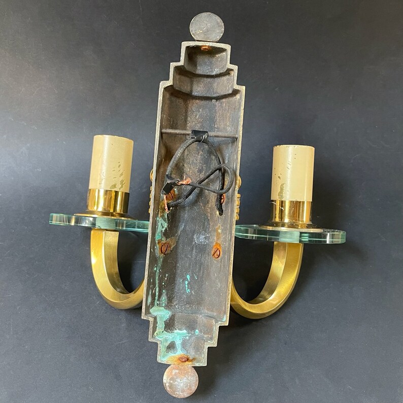 May include: Vintage brass wall sconce with two light fixtures. The sconce features a decorative design with clear glass accents and a central column. The sconce has a tarnished appearance, suggesting age. The sconce is designed to be mounted on a wall.