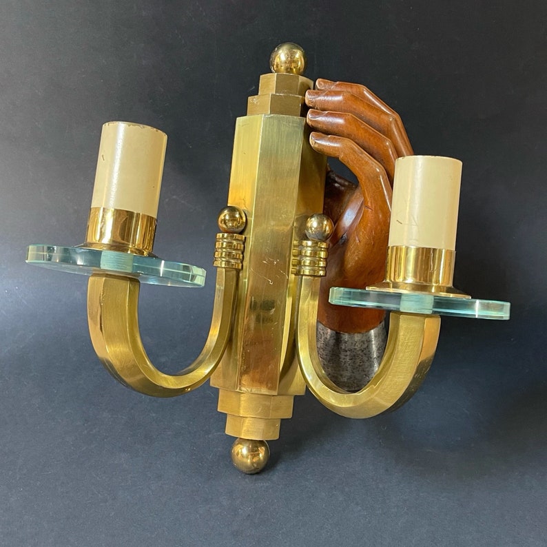 May include: Vintage brass wall sconce with two candle holders. The sconce features a stylized design with a wooden hand and clear glass accents. The sconce has a gold finish and is mounted on a dark background.