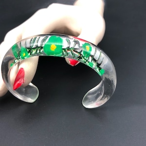 May include: A clear plastic bangle bracelet with a floral design. The flowers are green with yellow centers and are surrounded by a black and white pattern.