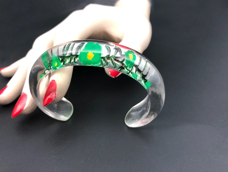 May include: A clear plastic bangle bracelet with a floral design. The flowers are green with yellow centers and are surrounded by a black and white pattern.