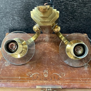 May include: A vintage brass wall sconce with two clear glass shades. The sconce has a geometric design and is mounted on a brown surface. The sconce has two light bulb sockets.