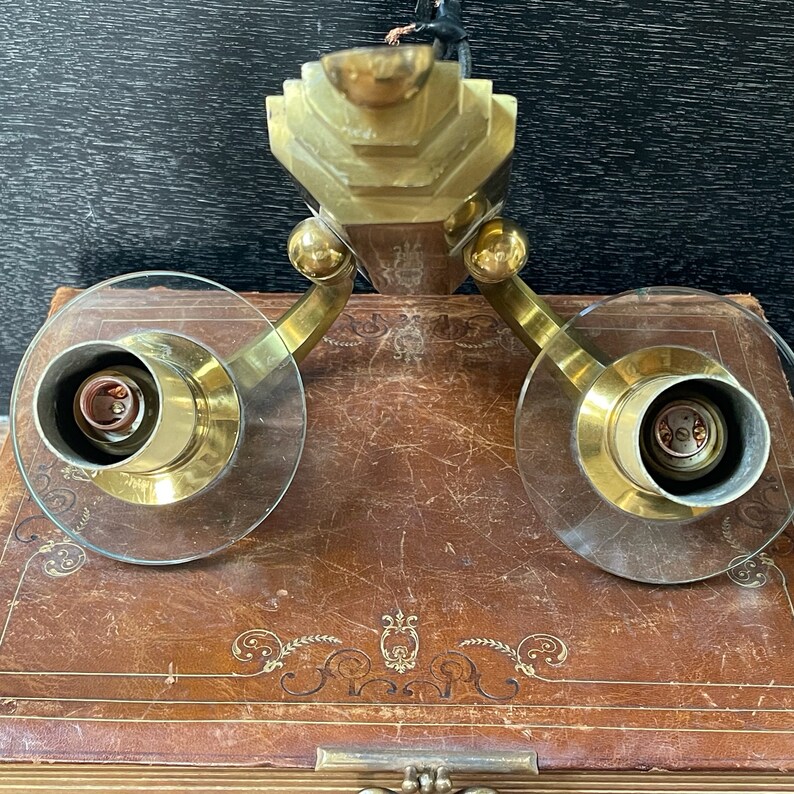 May include: A vintage brass wall sconce with two clear glass shades. The sconce has a geometric design and is mounted on a brown surface. The sconce has two light bulb sockets.