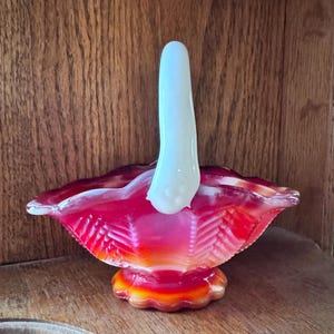 May include: A red, orange, and white glass candy dish with a white handle. The dish has a scalloped edge and a diamond pattern.