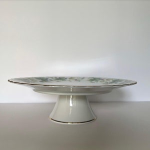 May include: White ceramic cake stand with a floral design and gold trim. The stand has a scalloped edge and a pedestal base.