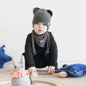 May include: A young child wearing a gray knitted hat with bear ears and a gray scarf sits on the floor. The child is looking at the camera. There are blue and white patterned pillows and a gray stuffed bunny on the floor.