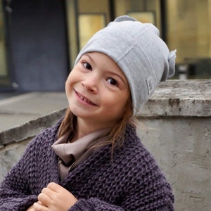 May include: A young girl wearing a gray knitted beanie with the word "btoo" on the side. She is smiling and wearing a dark purple knitted cardigan.