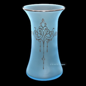 May include: A light blue glass vase with a silver rim and an intricate silver design. The vase has a flared top and a rounded bottom.
