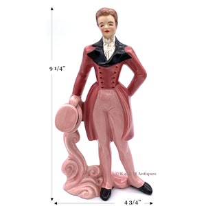 May include: A ceramic figurine of a person in a pink and black suit holding a hat. The figurine is 9 1/4 inches tall and 4 3/4 inches wide.