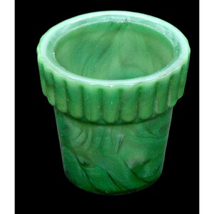 May include: A small, cylindrical green glass vessel featuring a marbled, swirling pattern throughout its body. The upper rim is defined by a distinct, vertical ribbed texture that encircles the top edge, creating a contrast with the smooth, flowing appearance of the base.