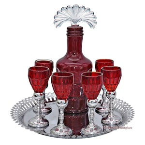 May include: A set of six ruby red glass shot glasses and a matching decanter with a clear glass stopper. The set is displayed on a silver tray with a decorative border.
