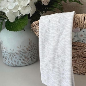 May include: A white headband with a delicate embossed pattern. The headband is lying in a wicker basket with a floral fabric lining.