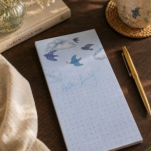 Può includere: Un blocco note azzurro con un motivo a griglia a pois e la scritta a mano "Hello, lovely!". Il blocco note presenta illustrazioni di uccelli e nuvole. Una penna d'oro è appoggiata accanto al blocco note su una superficie di legno.