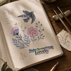 Könnte beinhalten: Ein offenes Notizbuch mit Blumen- und Vogelzeichnungen und den Worten "Make Something Lovely" auf einem Aufkleber. Das Notizbuch liegt auf einer Holzoberfläche, mit einer Tasse Tee und Kunstzubehör in der Nähe. Die Skizzen zeigen Blumen und einen Vogel.