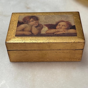 May include: A rectangular, gold-colored trinket box. The lid features a Renaissance-style painting of two cherubs with wings. The box has a decorative border and a slightly distressed finish, suggesting an antique or vintage aesthetic.